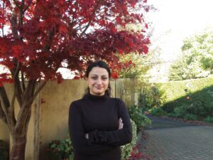 Woman standing in front of a brown fence next to tree with red leaves with arms folded wearing brown top and looking at the camera smiling.