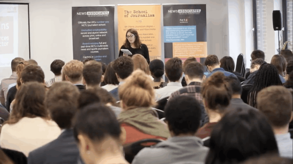 Woman stands in front of class of adults holding a note pad like she is presenting, background says school of journalism.