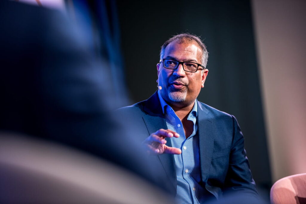 Asian man talking at conference wearing microphone and blue suit jacket with checked blue shirt and rectangle glasses.