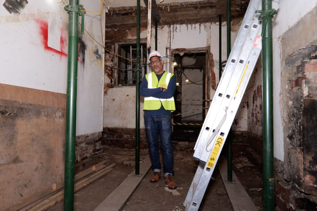 South Asian man standing with arms crossed wearing a high-vis vest and safety helmet, inside a construction site
